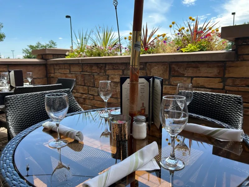 Outdoor table set with silverware and glasses, a drink menu, and an umbrella in front of a retaining wall
